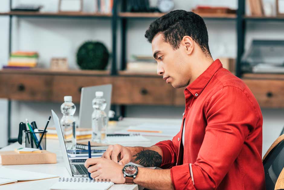 A man in a red shirt gazes at a computer screen in a working environment