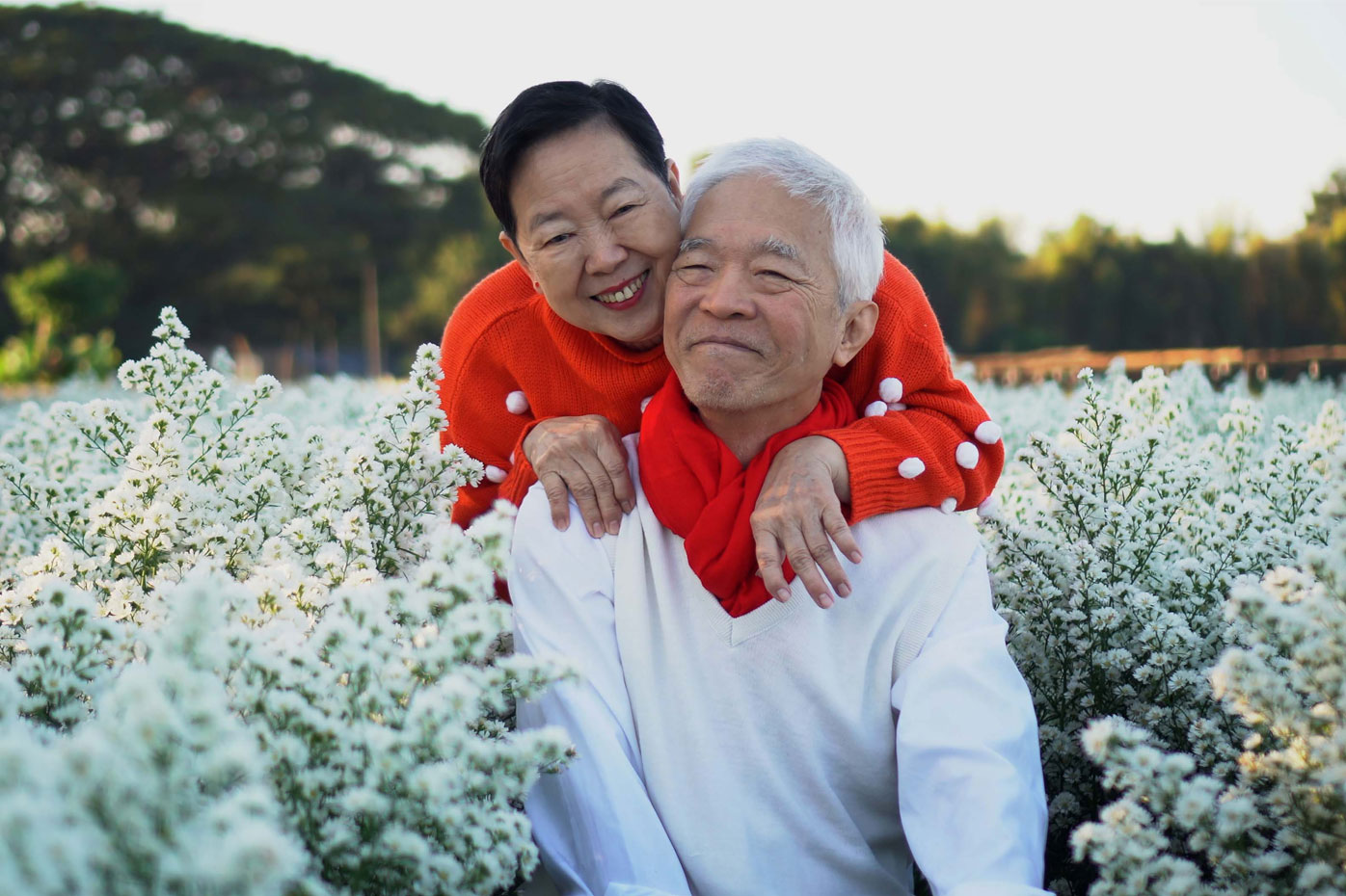 A smiling older couple of Asian descent sit in a field of white flowers