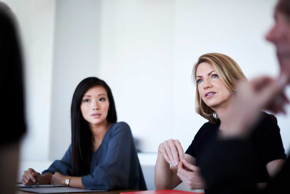 A pair of women engage in serious conversation around a business table.